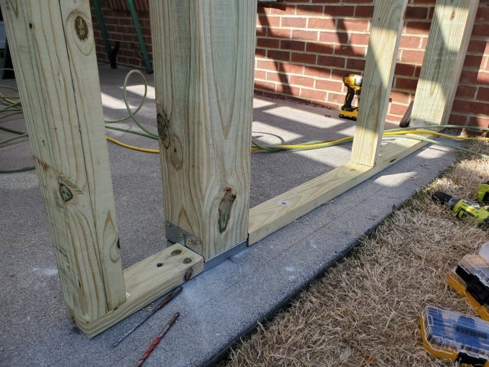 Wooden deck framing posts and beams beside a house, with concrete footers and a brick wall in the background.