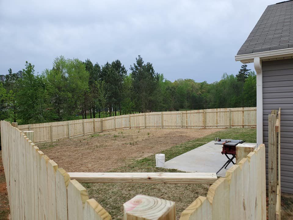 Wooden-fenced backyard construction area beside a house, with dirt ground and a concrete patio slab