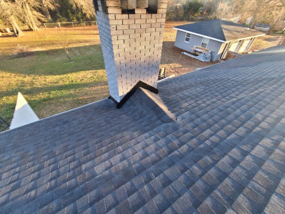 Rooftop view of gray asphalt shingles around a white brick chimney in a suburban yard.