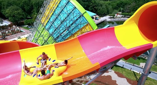 Two people on a bright yellow, pink, and orange water slide, splashing down in a lush outdoor park.