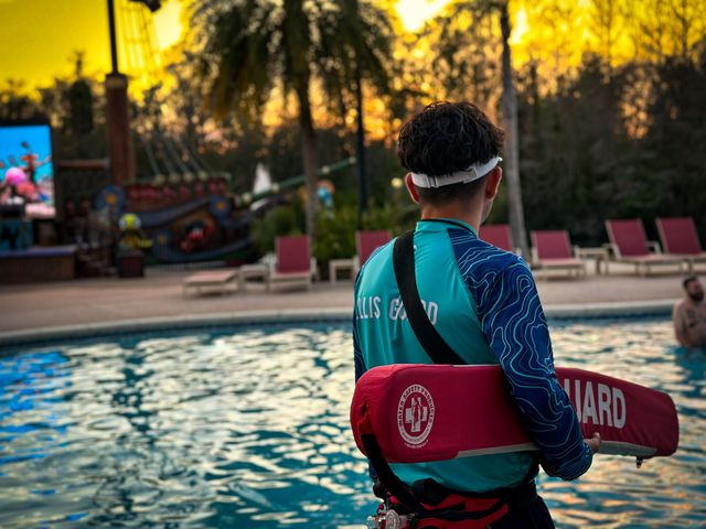 Lifeguard in teal uniform holding a red rescue float beside a pool at sunset
