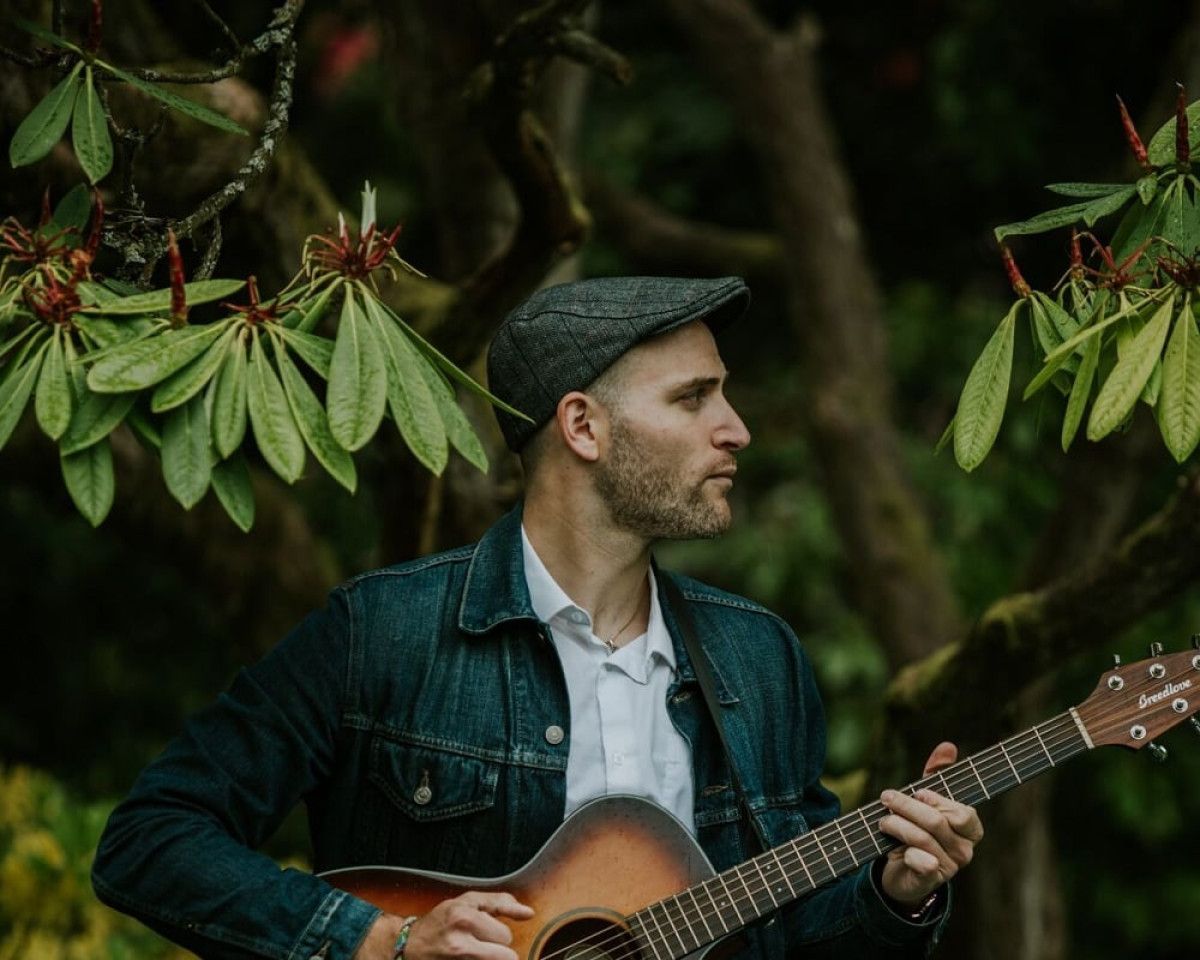 Jeremy Levif, wearing a button down shirt and holding a guitar, sings into a microphone. The background is black with a blue spotlight behind him.