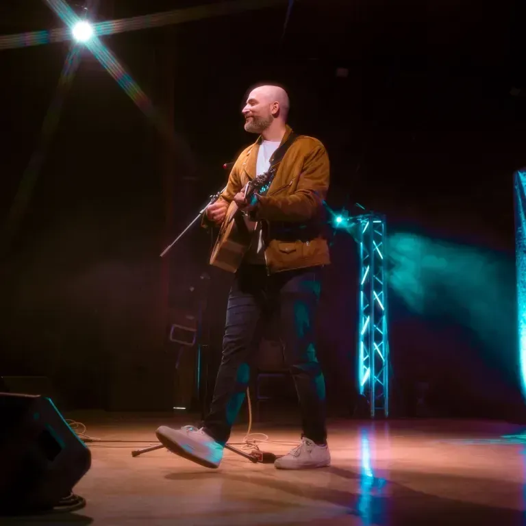 Jeremy Levif, wearing a button down shirt and holding a guitar, sings into a microphone. The background is black with a blue spotlight behind him.