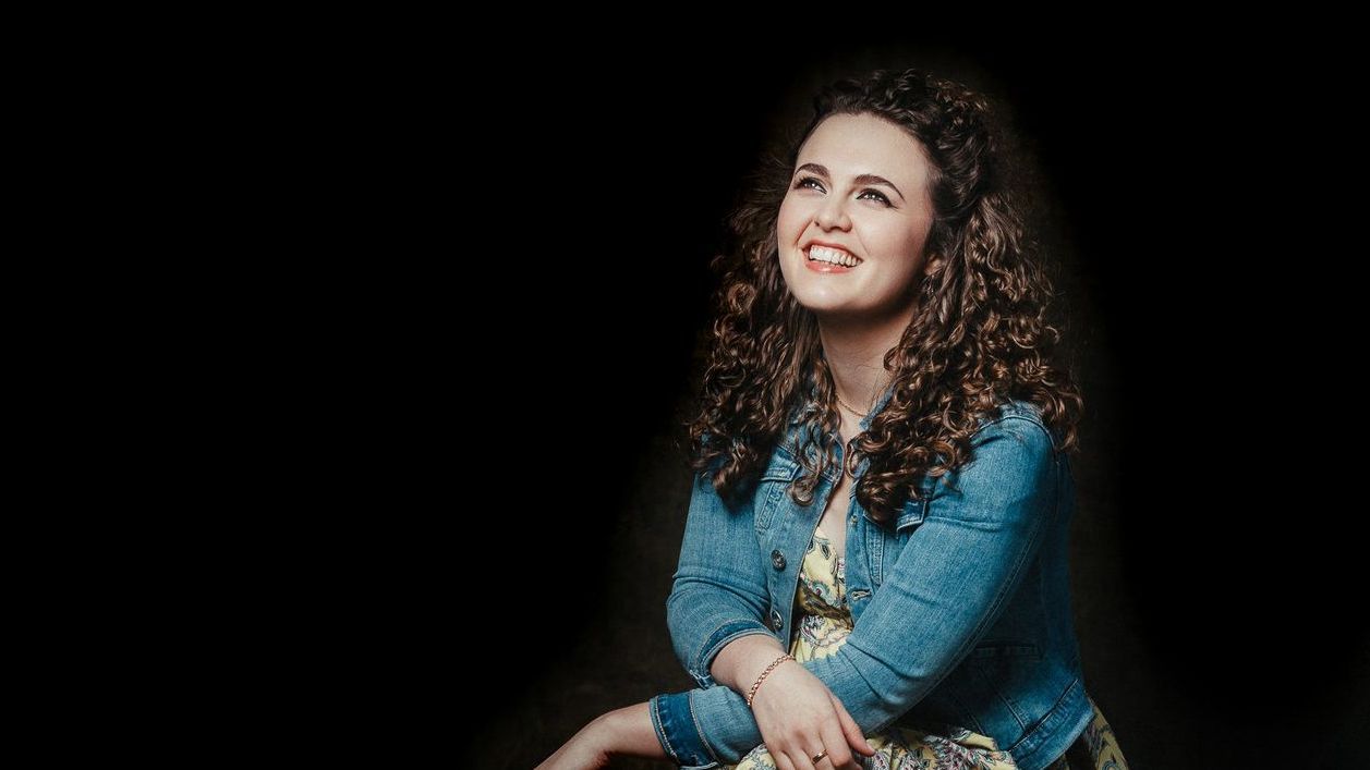 Katee Kross sitting smiling - she wears a denim jacket and has curly hair