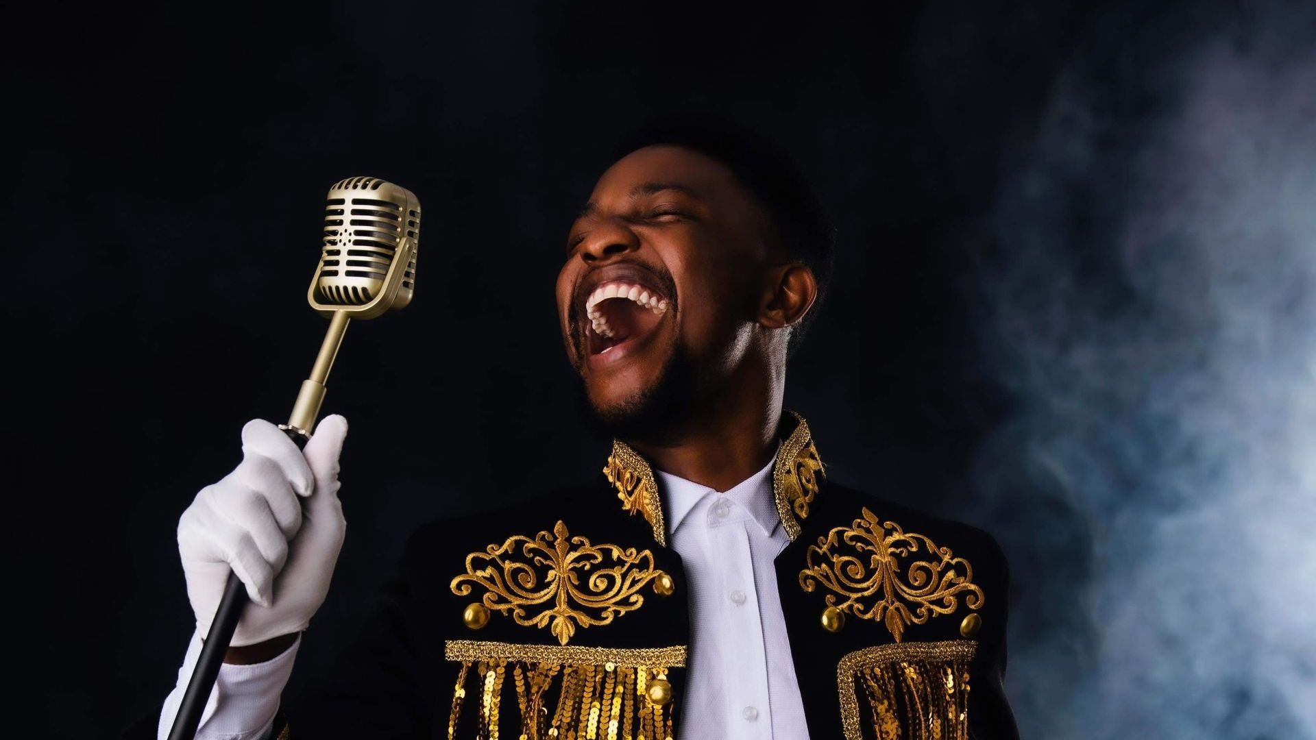 Simon Sims, wearing a black and gold jacket, sings into a microphone. The background is black with s