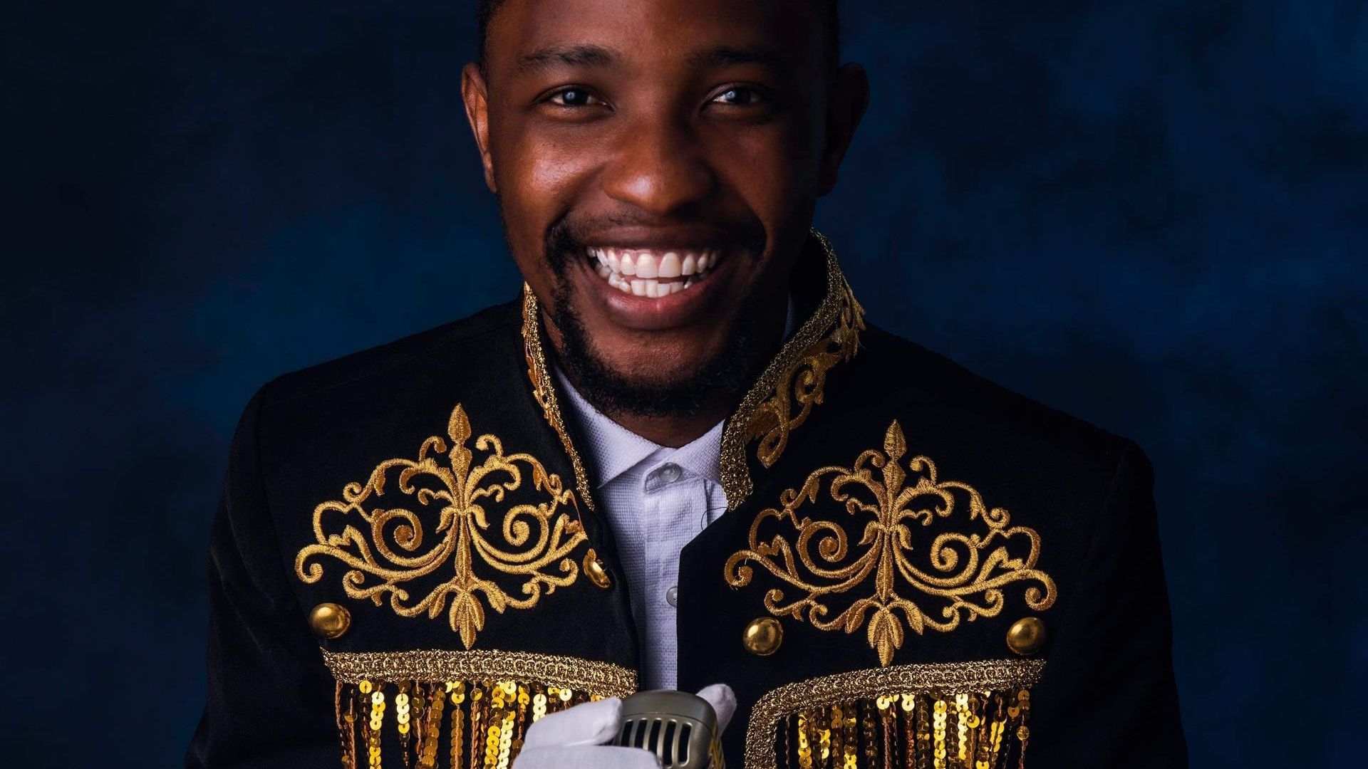 Simon Sims, a male singer, smiling into the camera. He is wearing a white shirt and gloves, and a black and gold jacket. He is holding a microphone. 