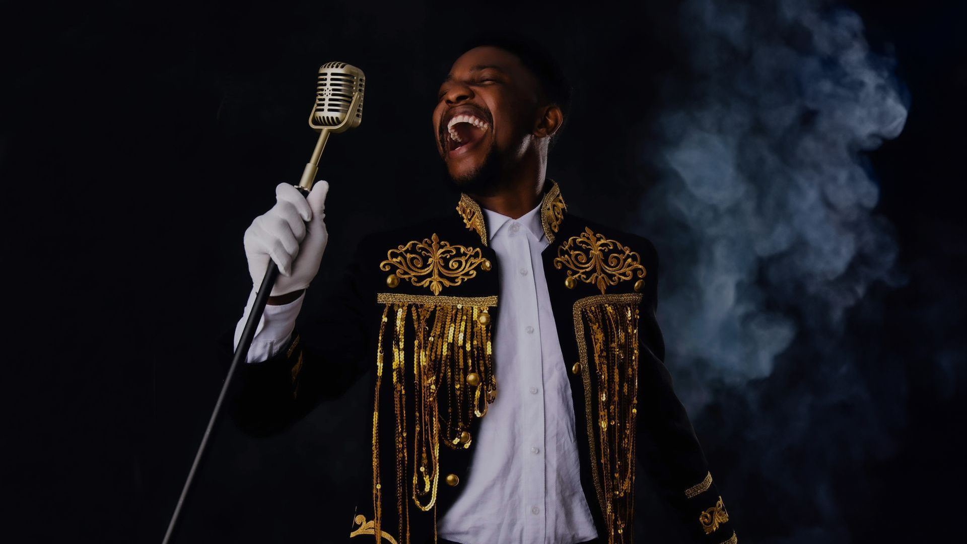 Simon Sims, a male singer, singing into a gold microphone. Simon wears a white shirt with a black and gold jacket, and white gloves. The background of the photo is black with some grey smoke.