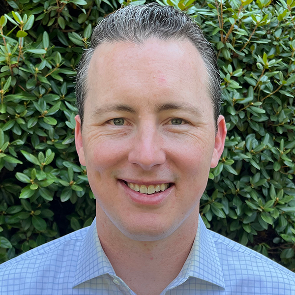 Smiling person in a light blue shirt standing in front of green bushes