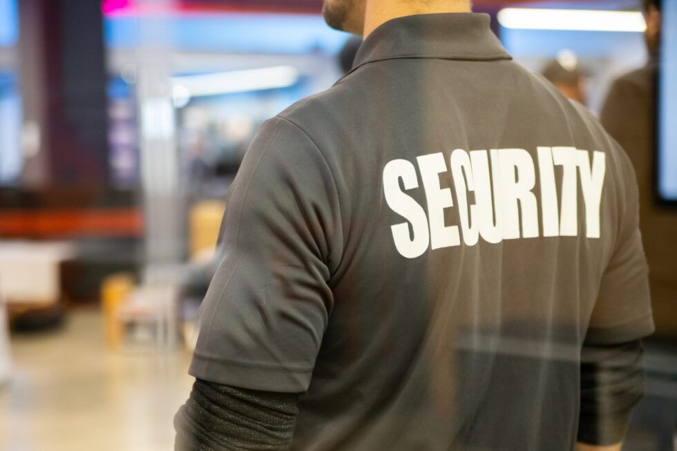 Security guard in dark jacket with large white “SECURITY” text, seen from behind indoors.