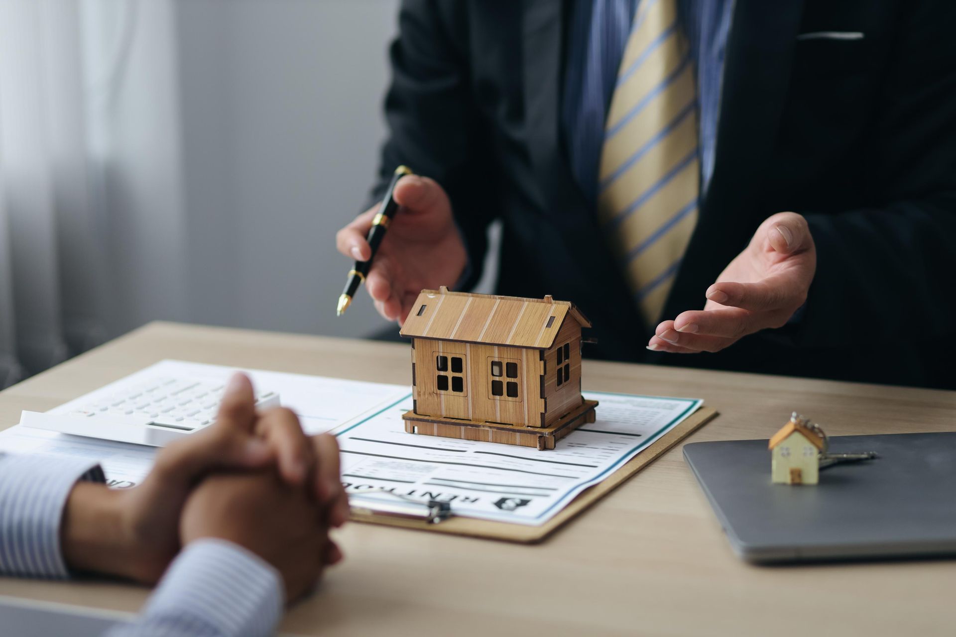 Real estate agent discussing house plans with a client; wooden model house on paperwork.