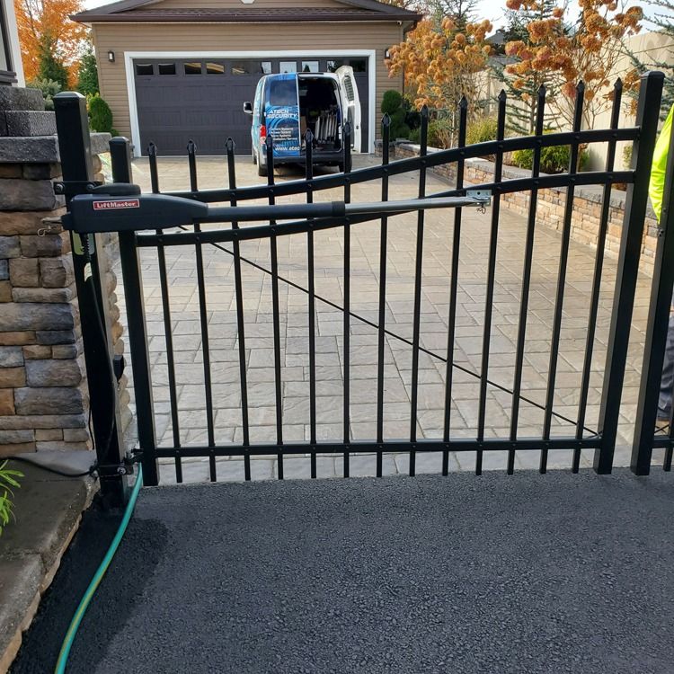 Black metal driveway gate at the entrance to a paved driveway with a house and van beyond.