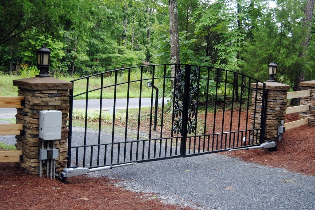 Black wrought-iron gate between stone pillars at a wooded gravel driveway entrance.