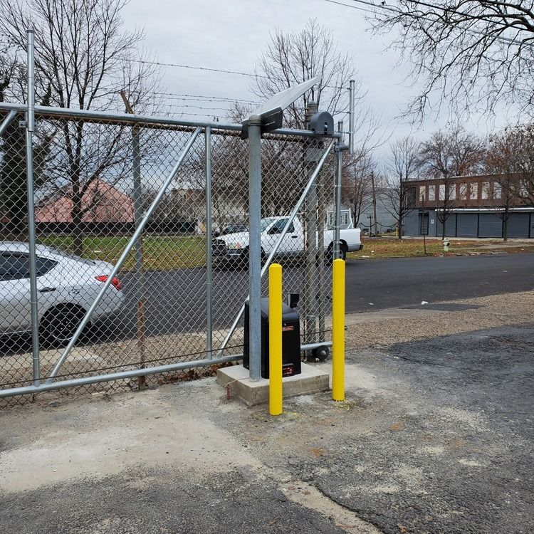 Chain-link gate with a black access control box and yellow bollards beside a parking lot