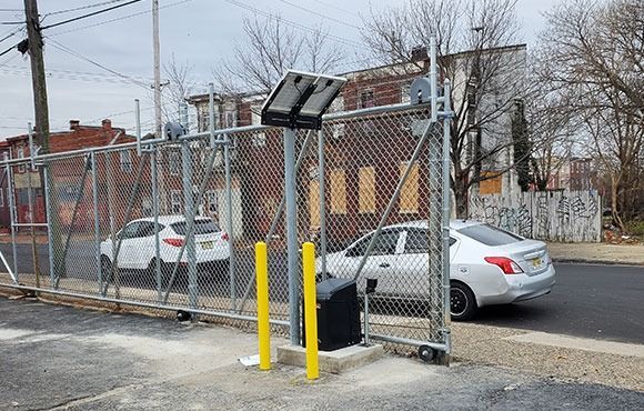 Chain-link gate with yellow bollards beside a street, with parked cars and brick buildings behind it.