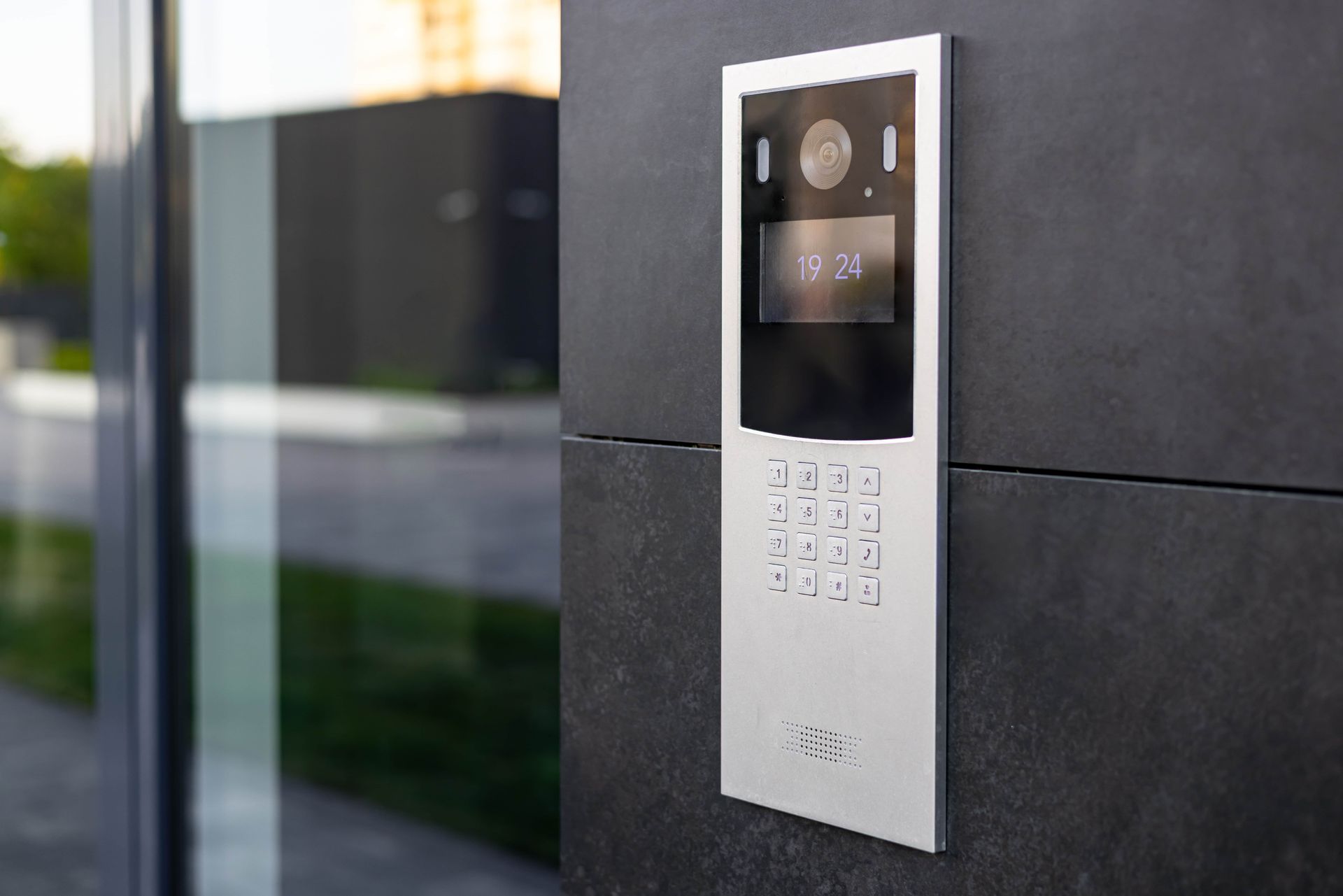 Silver video intercom panel mounted on a dark wall beside a glass door