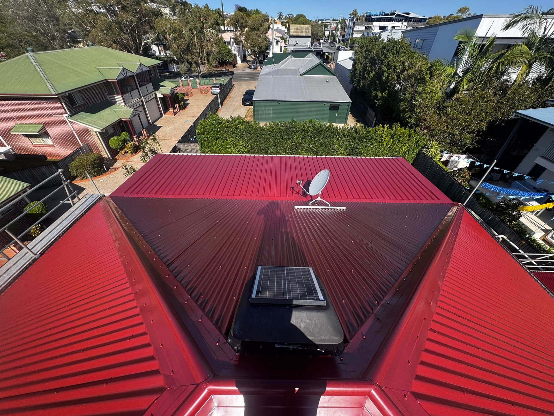 Red corrugated metal roof with solar panel and satellite dish, overlooking a residential neighborhood — Rumara Roofing in Sunshine Coast, QLD