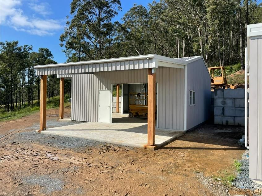 A Grey Corrugated Metal Shed With a Carport, Supported by Wooden Posts, on a Gravel Lot, With a Hillside in the Background — Bridgey's Constructions in Forster, NSW