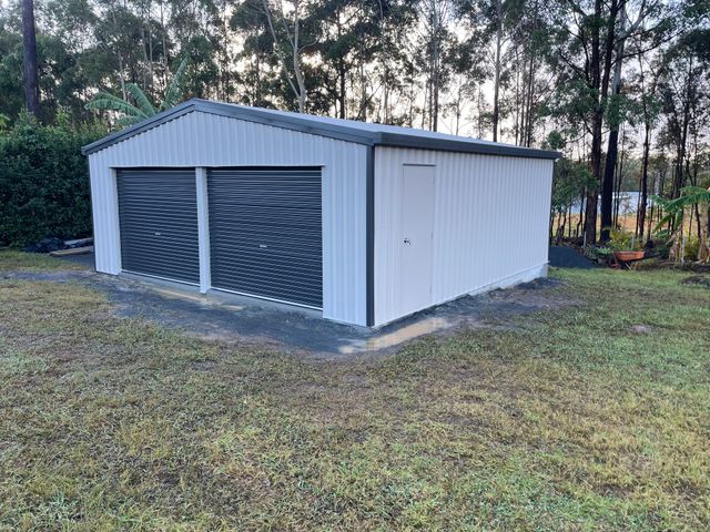 Small White Metal Shed with Blue Roof and Dark Roll Up Doors — Bridgey's Constructions in Wallabi Point, NSW