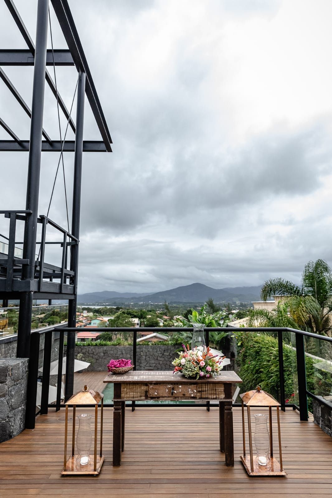 Terraço com varanda, mesa de madeira, lanternas e flores, com vista para a paisagem montanhosa encoberta por nuvens.