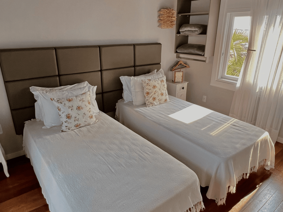 Quarto luminoso com duas camas de solteiro, roupa de cama branca, piso de madeira e luz solar entrando pela janela.