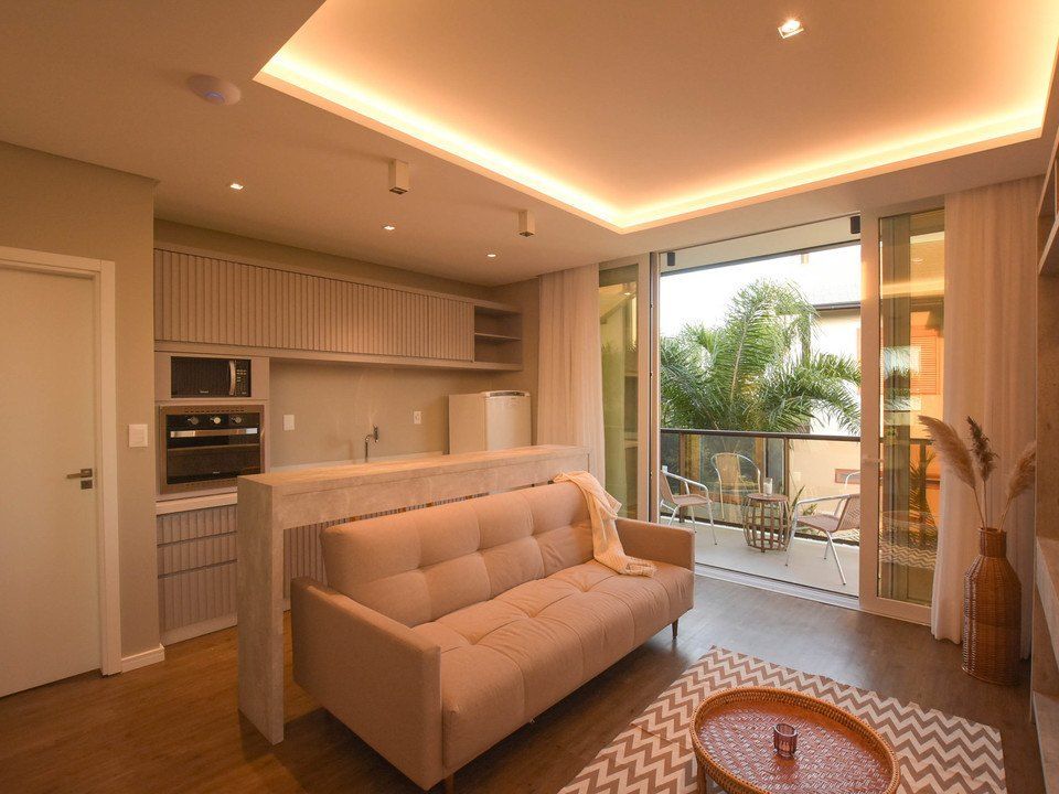 Sala de estar moderna com sofá bege, cozinha compacta, portas para varanda e iluminação aconchegante no teto.