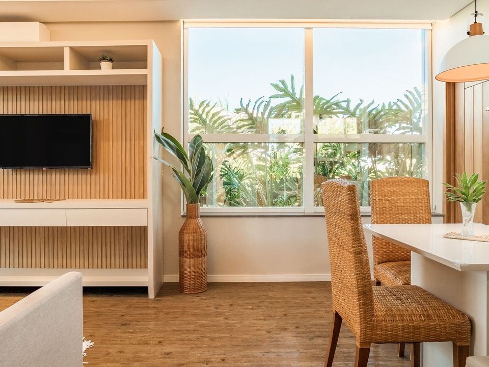 Sala de estar iluminada com TV, janela grande, plantas e cadeiras de jantar de vime.
