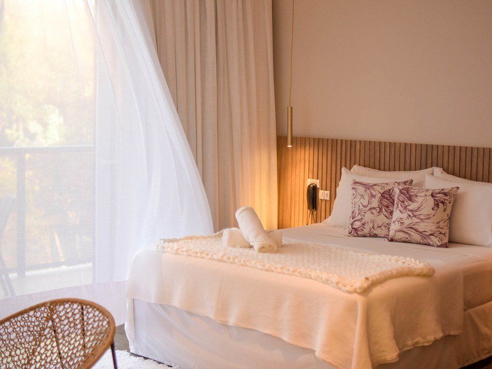 Quarto luminoso com cama, cortinas translúcidas, abajur aconchegante e uma cadeira de vime junto à janela.