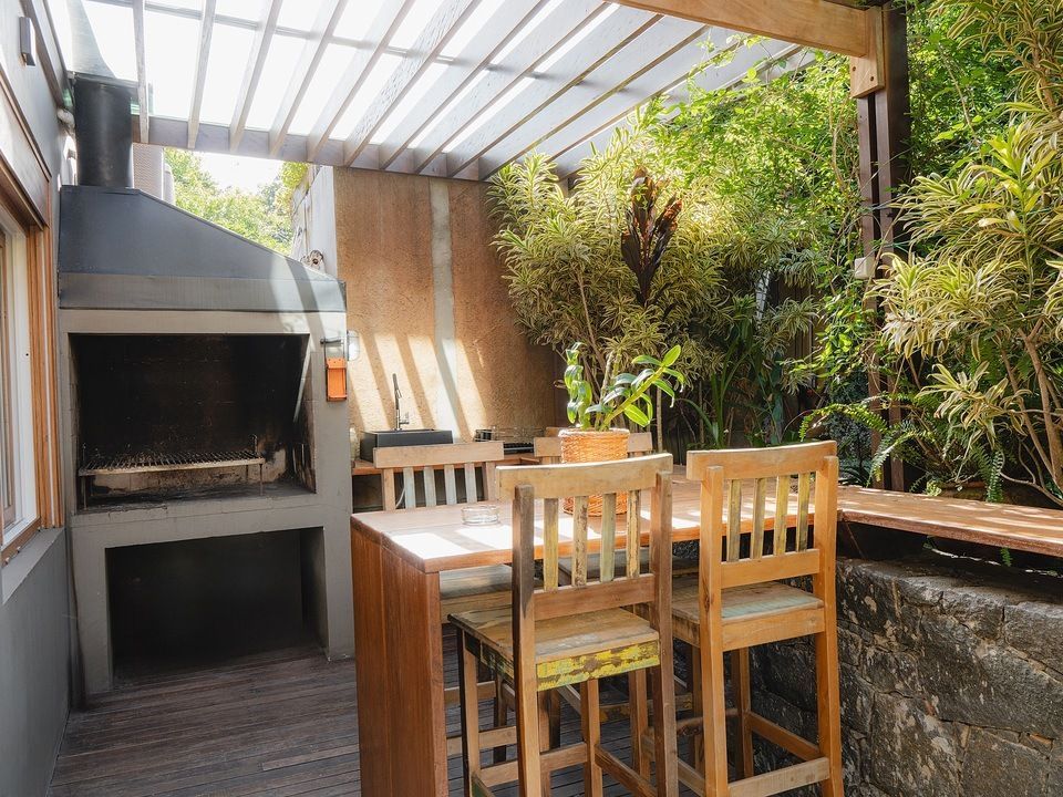 Pátio externo com mesa de jantar de madeira, bancos e churrasqueira embutida, sob cobertura e rodeado de plantas.