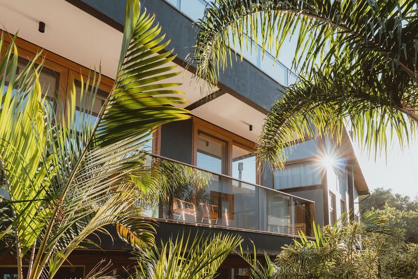 Prédio de apartamentos moderno com varandas de vidro e palmeiras, banhado por luz solar.