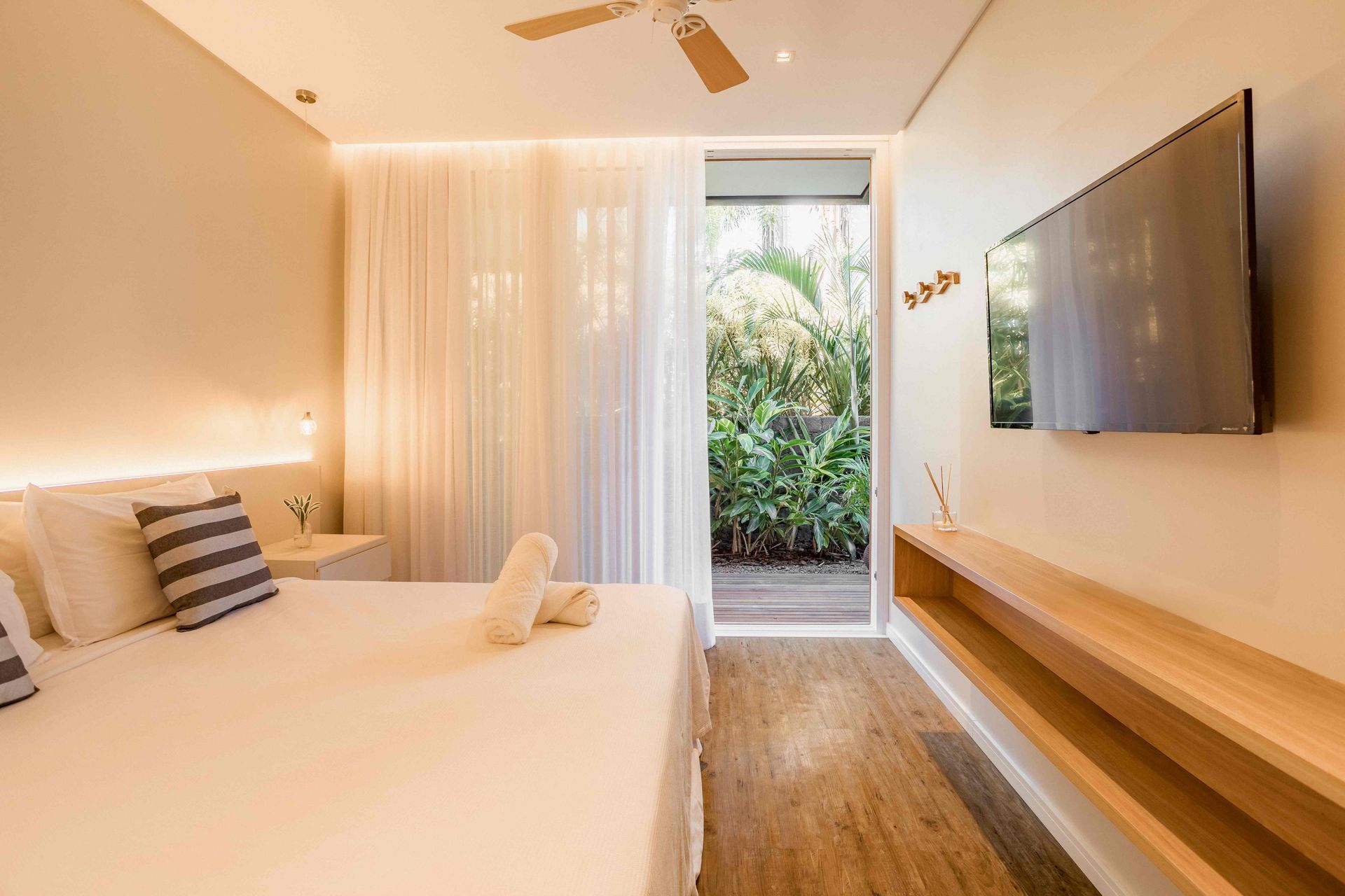 Quarto minimalista e luminoso com cama, TV de parede e porta aberta para um pátio verde.