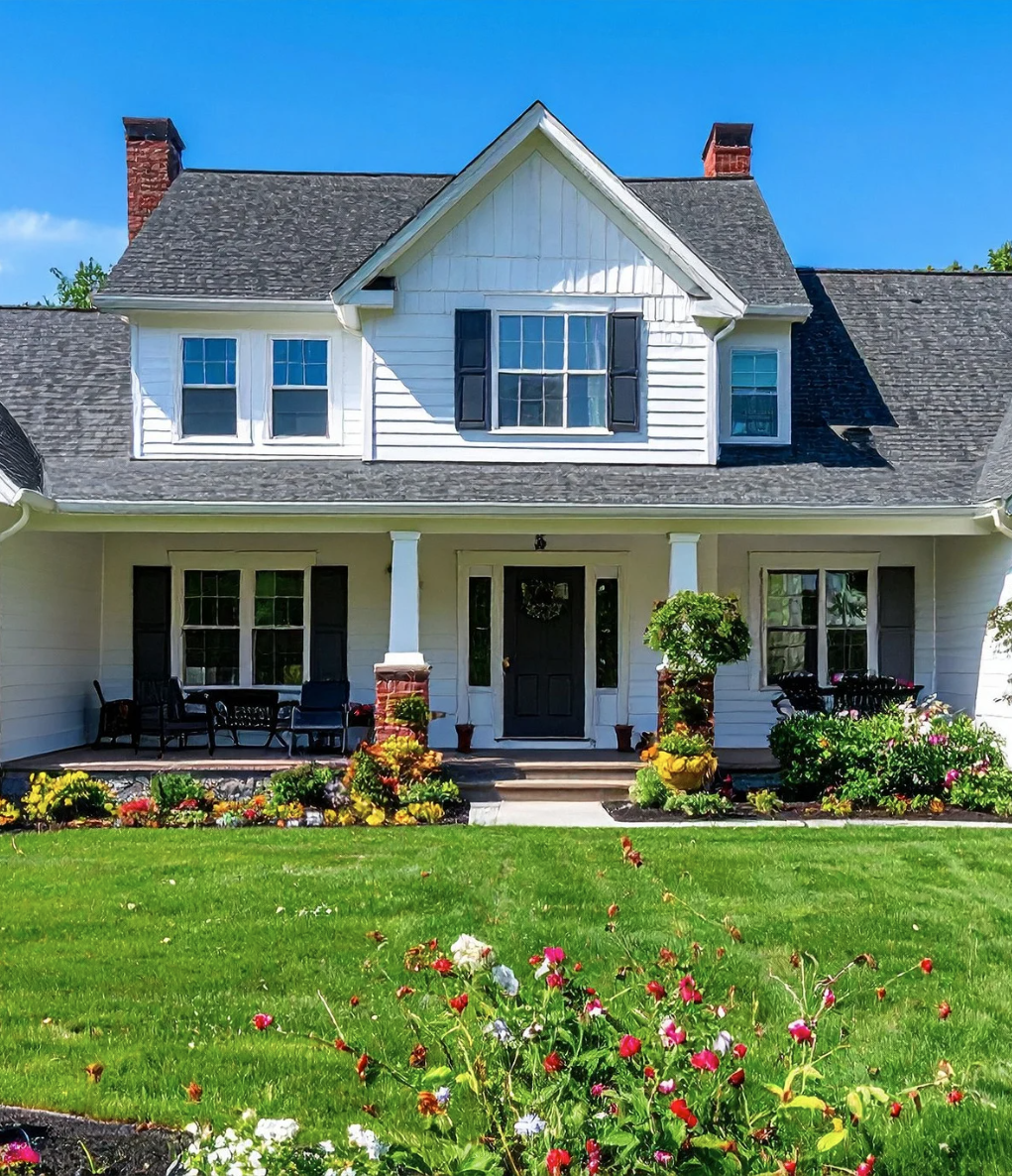 White farmhouse with black shutters and a front porch, surrounded by a green lawn and flowers under a blue sky