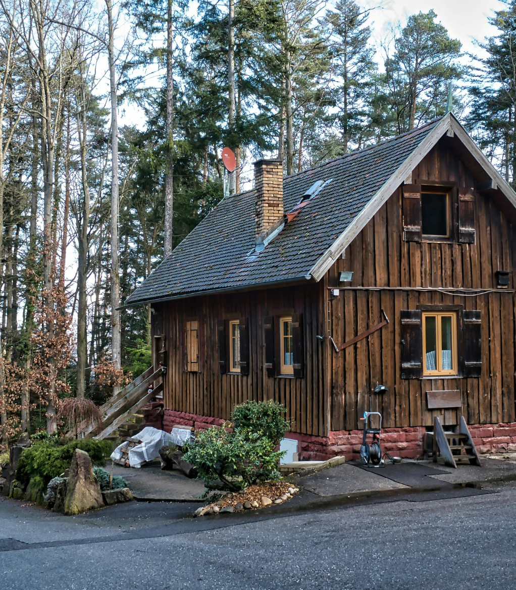 Rustic wooden cabin in a forest, with a steep roof and chimney beside a paved road.