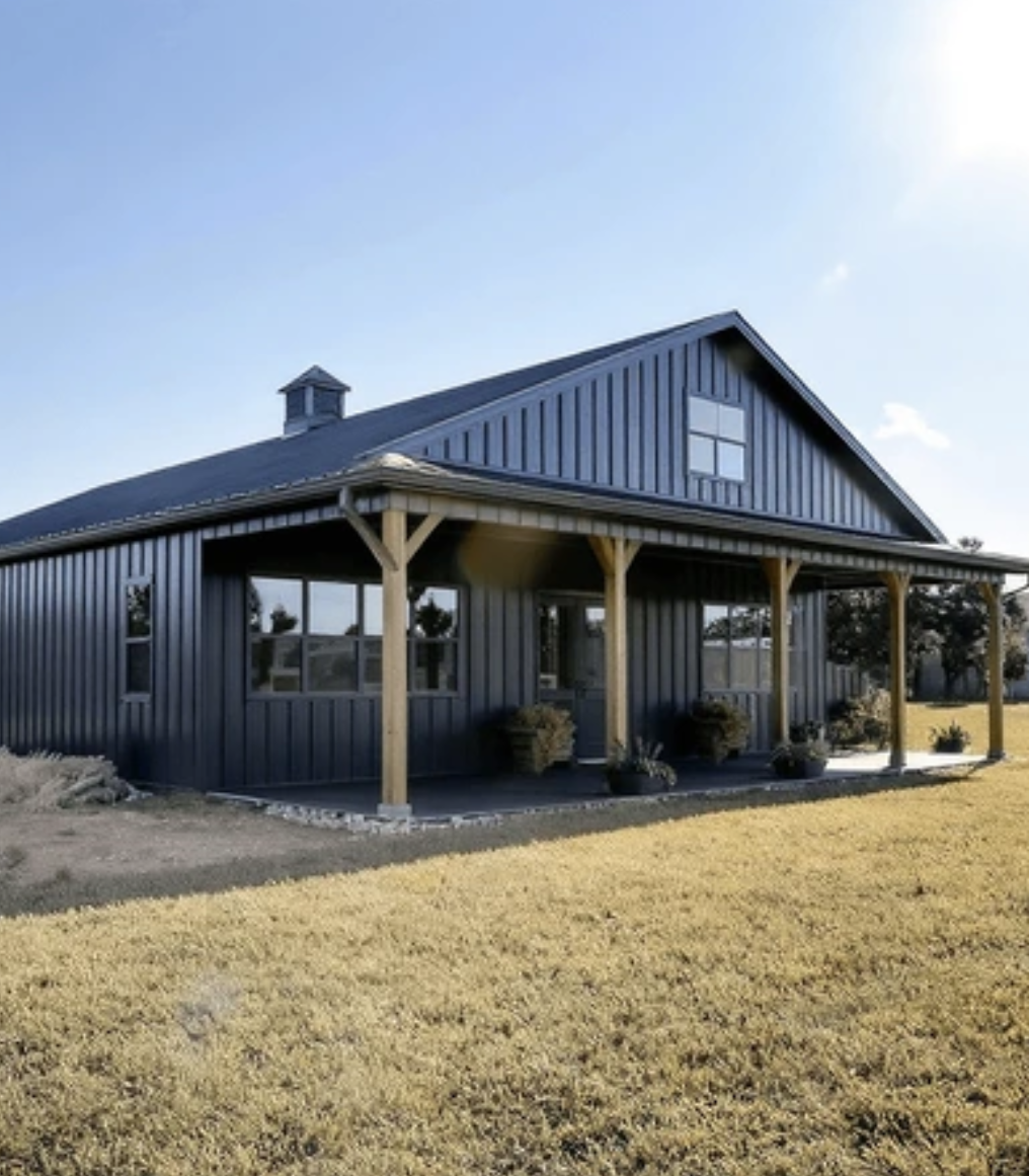 Dark gray metal barn-style house with a covered porch and dormer, set in a grassy field under a blue sky