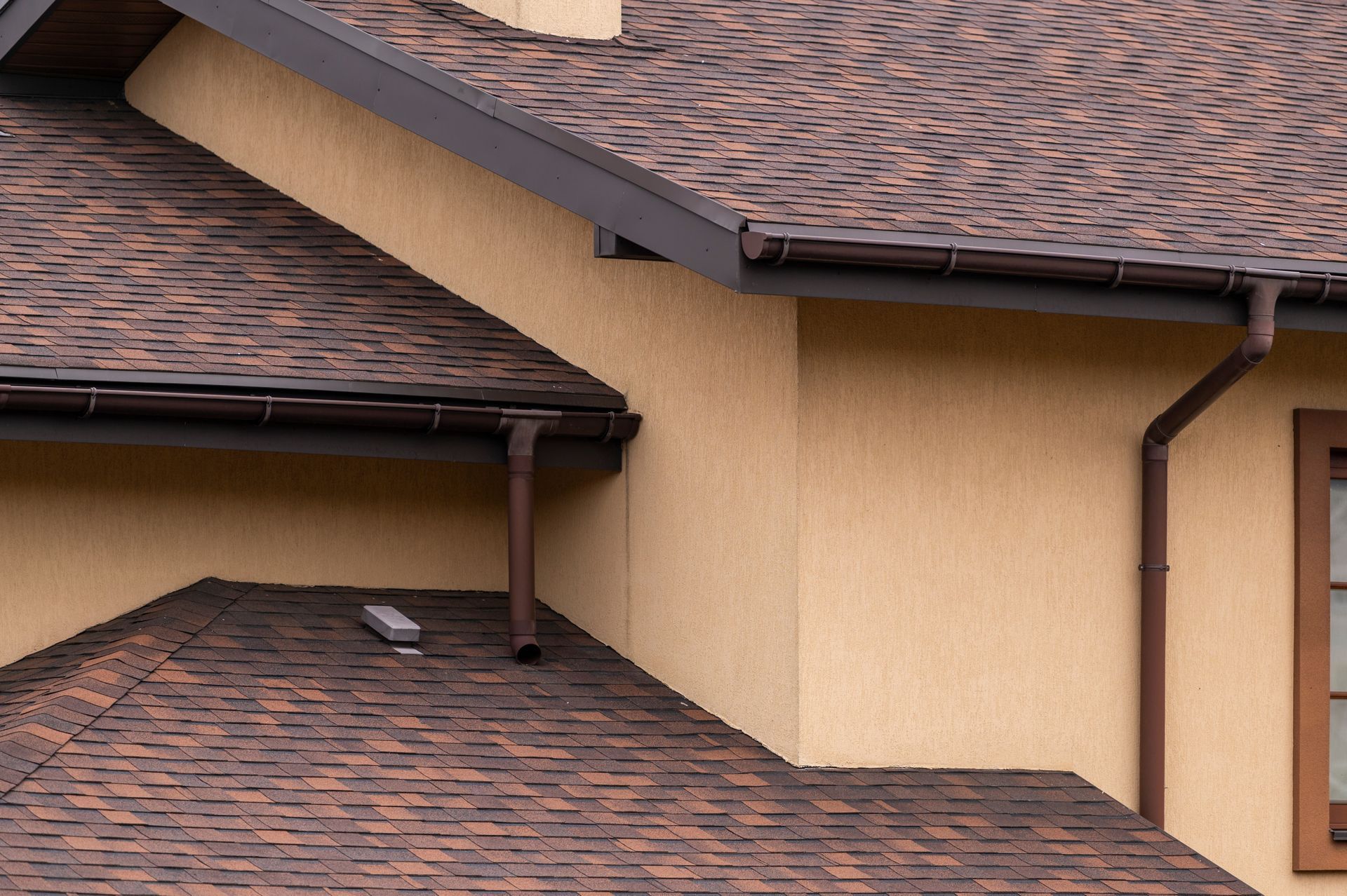 Close-up of a tan house with brown shingle roofs and dark gutters and downspouts.
