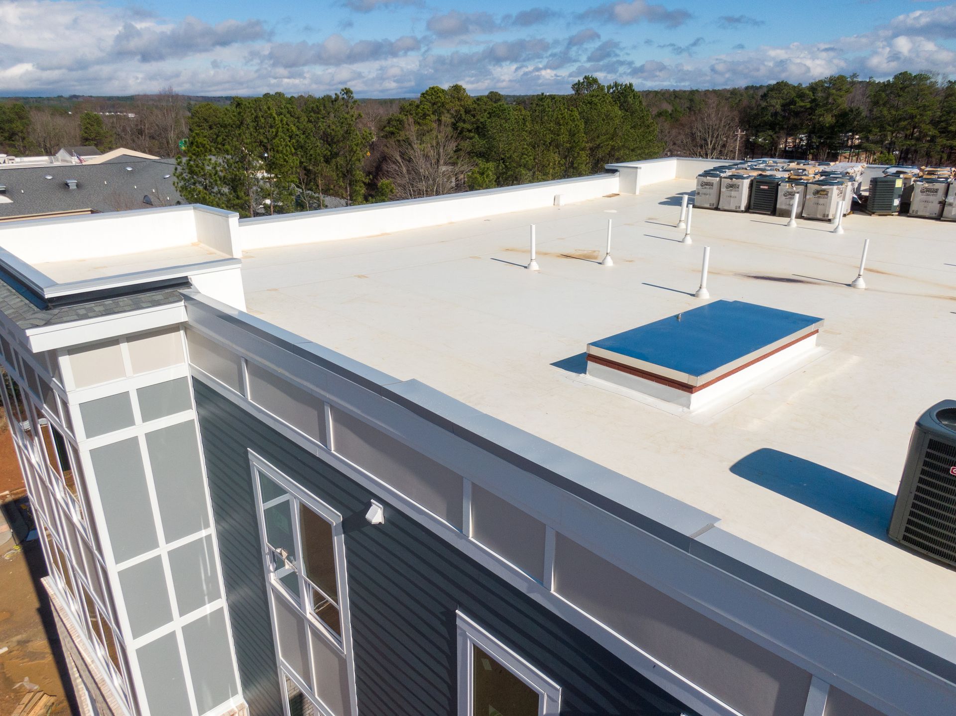 Rooftop view of a flat white roof with HVAC units, skylight, and surrounding trees under a cloudy sky