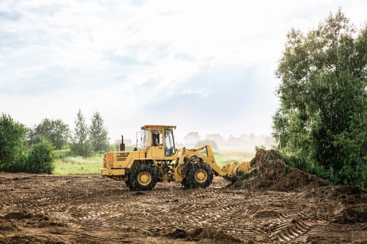 Yellow bulldozer clearing dirt in a field beside trees under a cloudy sky