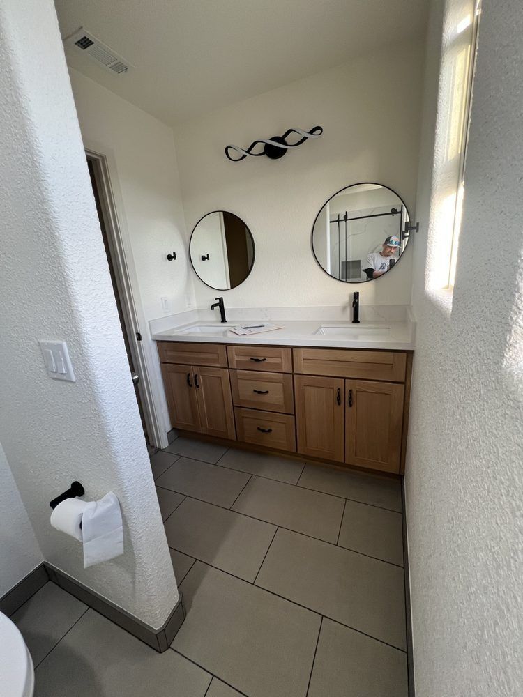 Small bathroom with wooden vanity, double round mirrors, white walls, and tiled floor.
