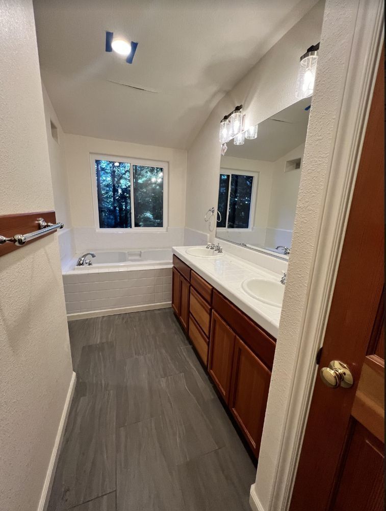 Bright bathroom with double vanity, large mirror, gray floor, and a window above a bathtub.