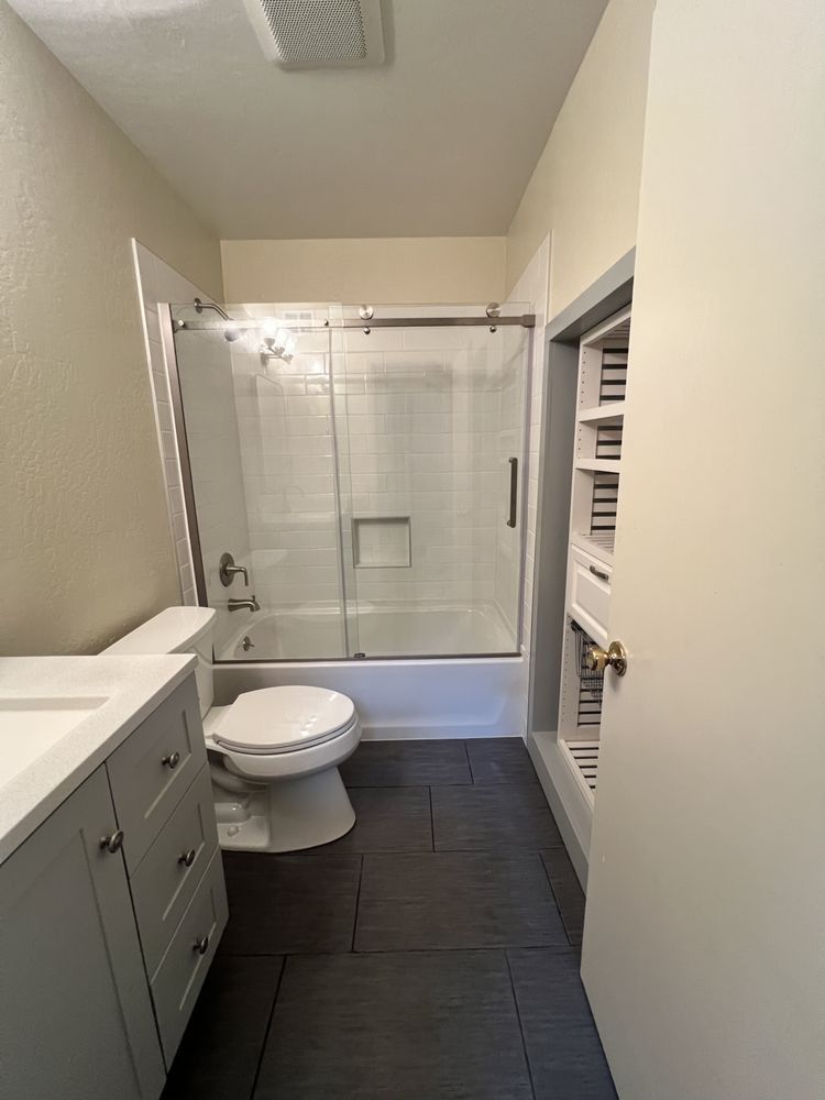 Small bathroom with white vanity, toilet, dark tile floor, and glass-enclosed bathtub shower.