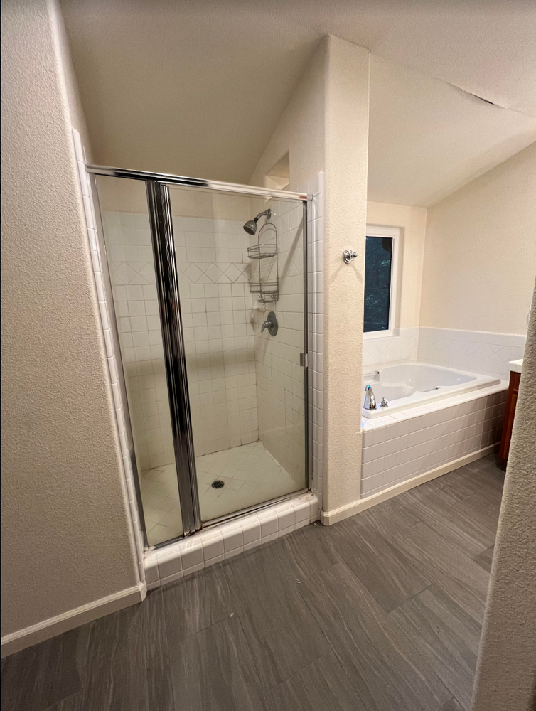 Bathroom with glass shower stall, white bathtub, and gray wood-look flooring.