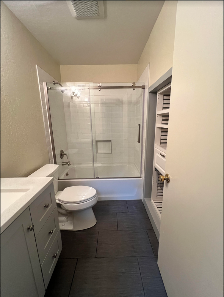 Small bathroom with gray vanity, toilet, glass shower, and dark tile floor.