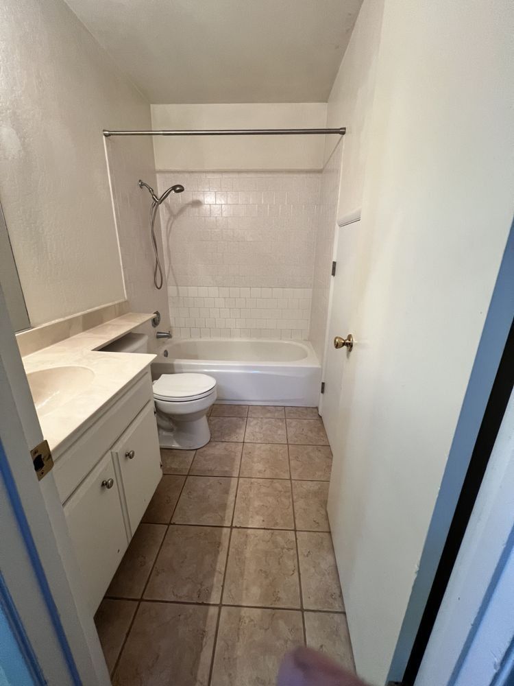 Narrow bathroom with white vanity, toilet, tub, and beige tile floor viewed from the doorway
