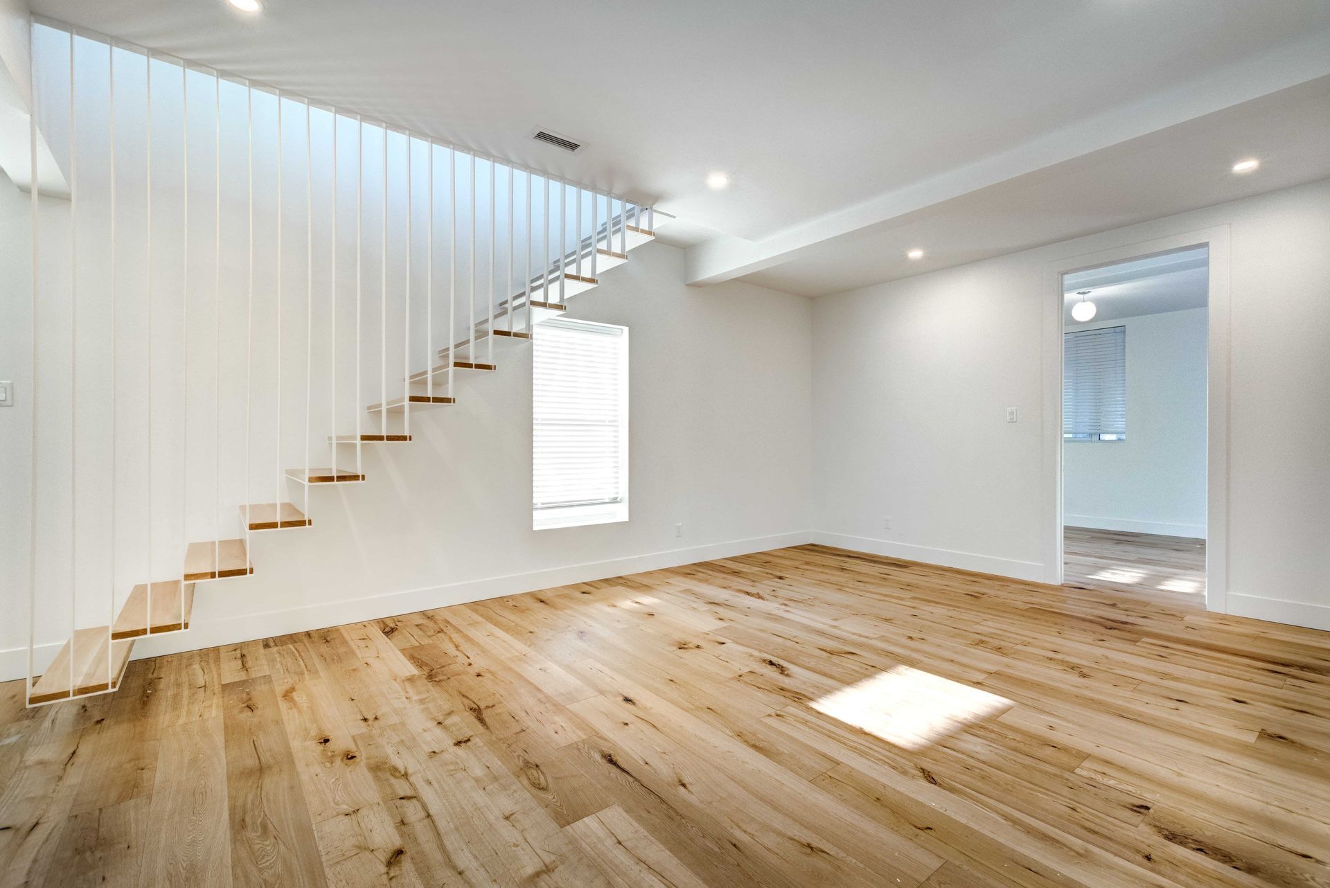 Bright empty room with wood floors, white walls, and floating staircase by tall windows