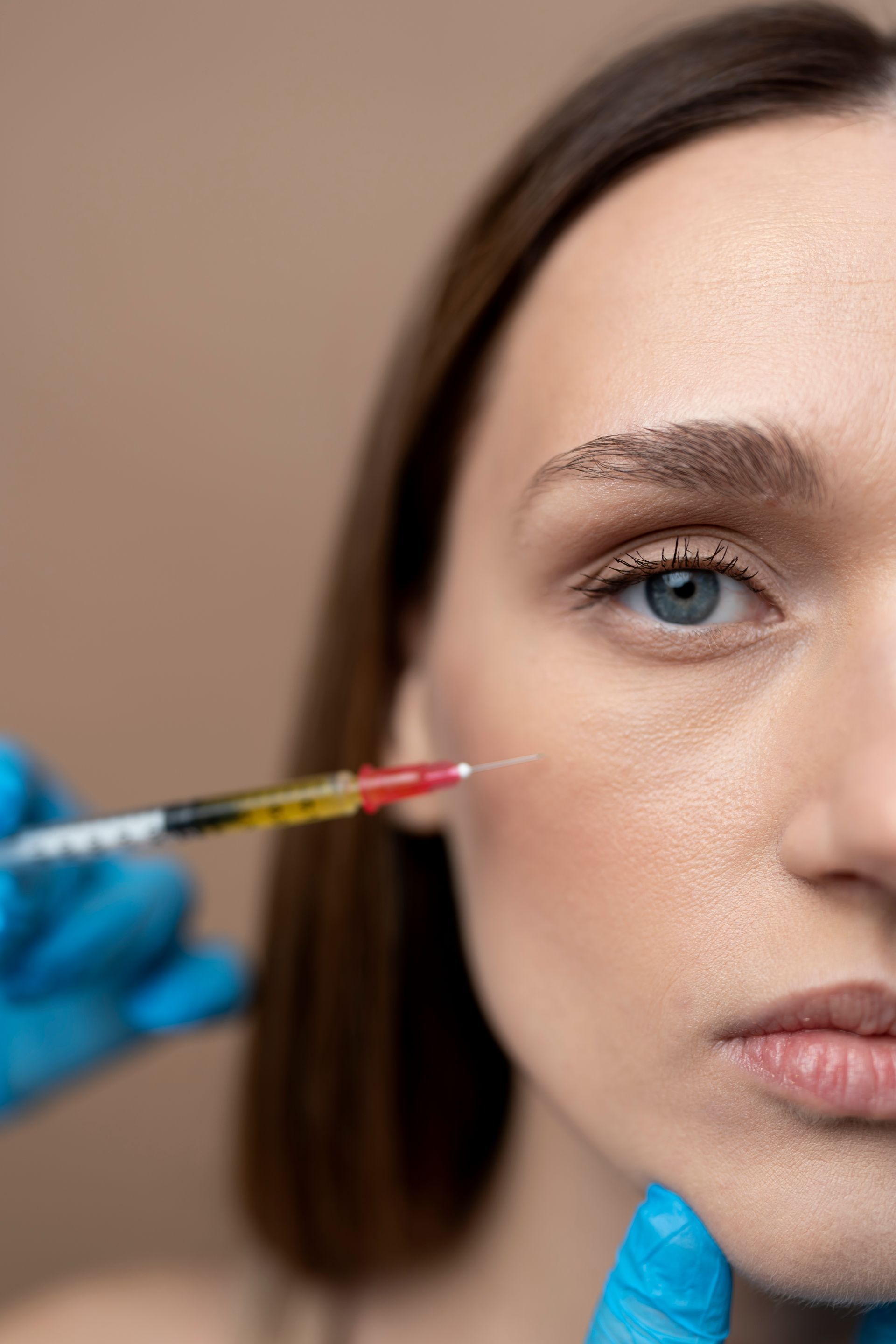 Woman receiving cosmetic injection near cheekbone; blue gloves, syringe, neutral background.