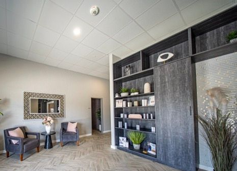 Modern waiting area with two armchairs, wall mirror, and dark built-in shelving beside a glass door.