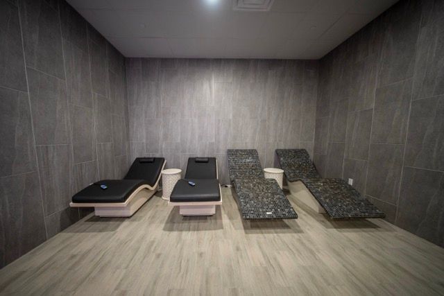 Spa room with four cushioned lounge chairs lined up against gray tiled walls and a wooden floor.