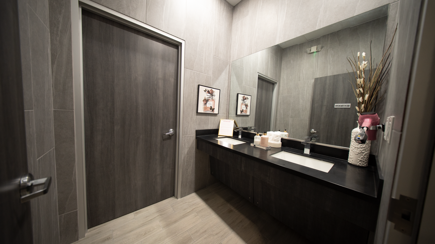 Modern restroom with dark wood stalls, gray walls, a black sink counter, and a vase of flowers.