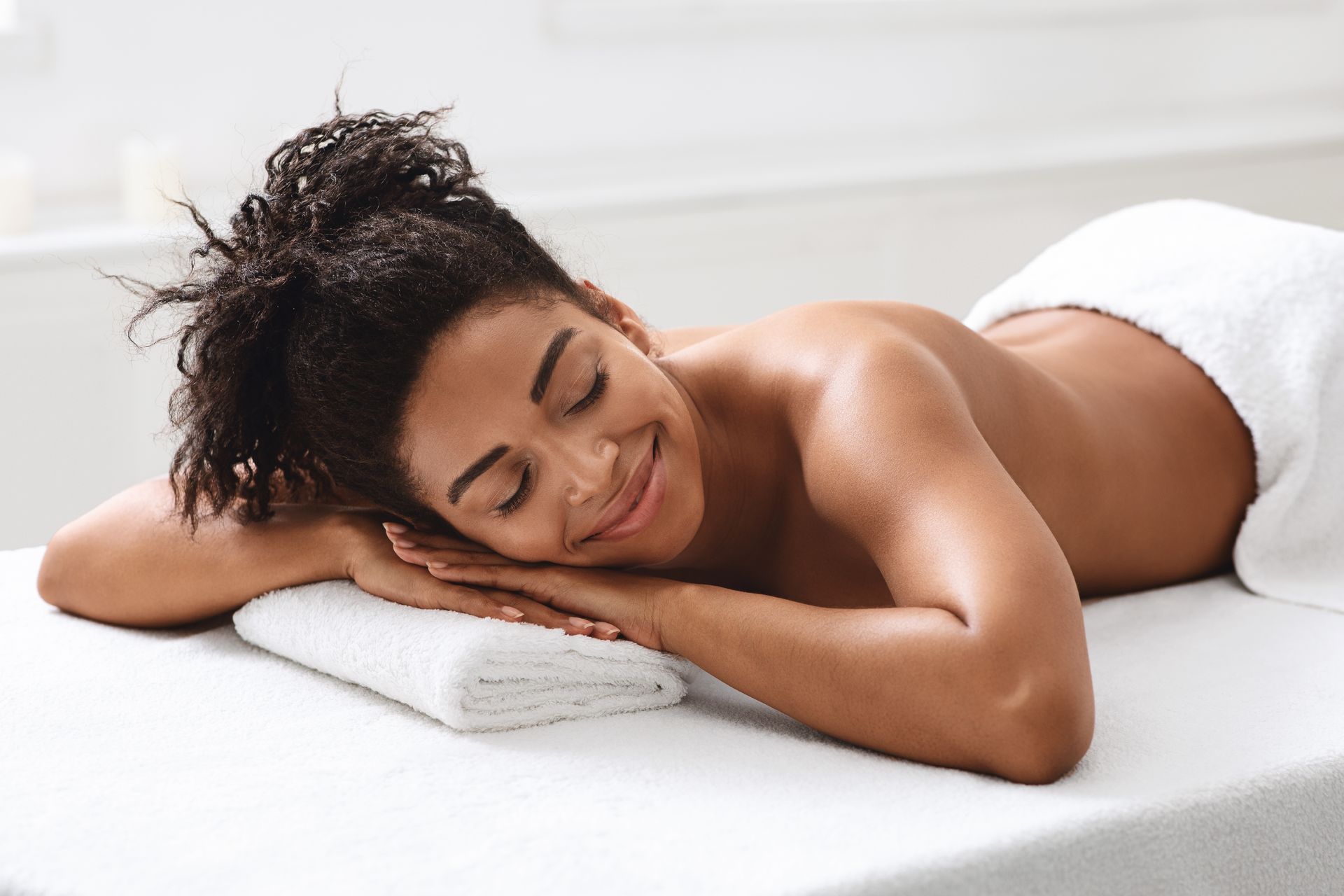 Woman relaxed on massage table, eyes closed, smiling, white towel, spa setting.