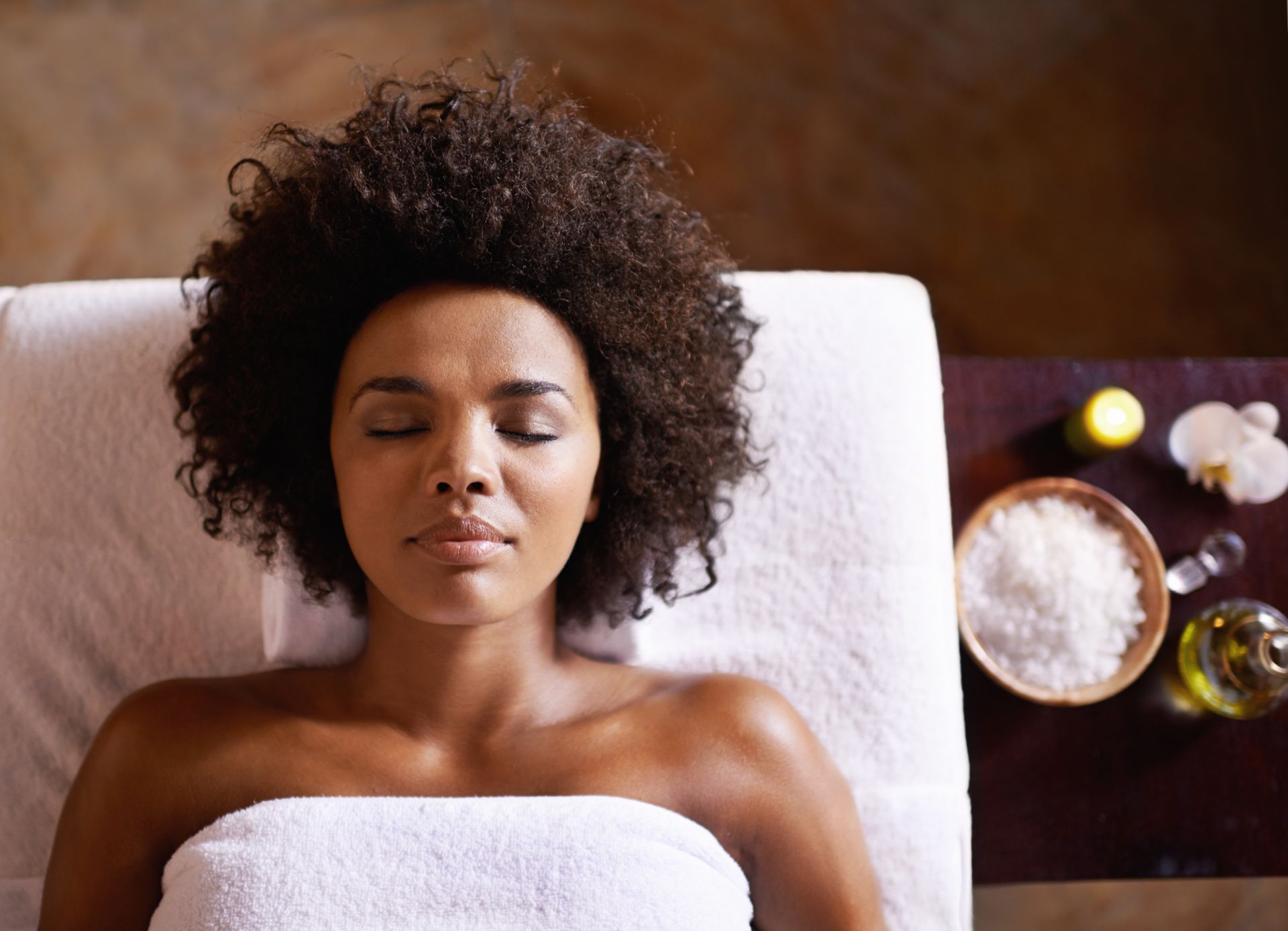 Woman receiving a spa facial treatment, eyes closed, lying on a towel with skincare products nearby
