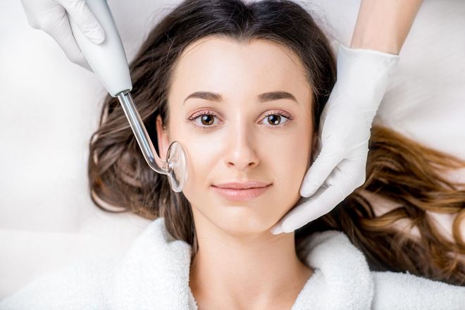 Woman receiving a facial treatment with a handheld device and gloved hands on a spa bed